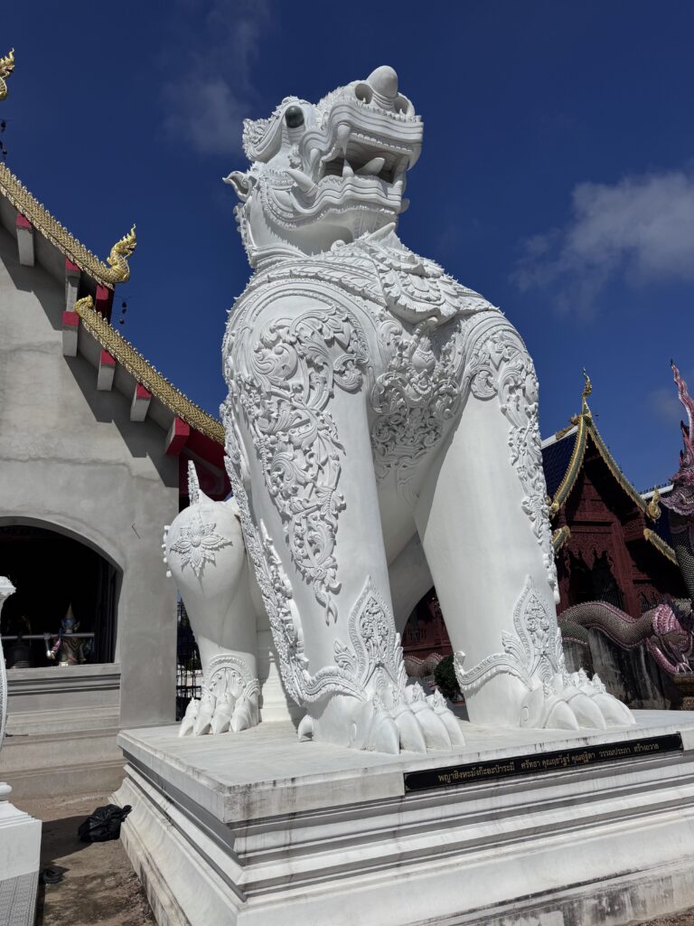 White, intricately carved lion statue stands guard outside a temple, showcasing traditional Thai architecture against a clear blue sky.