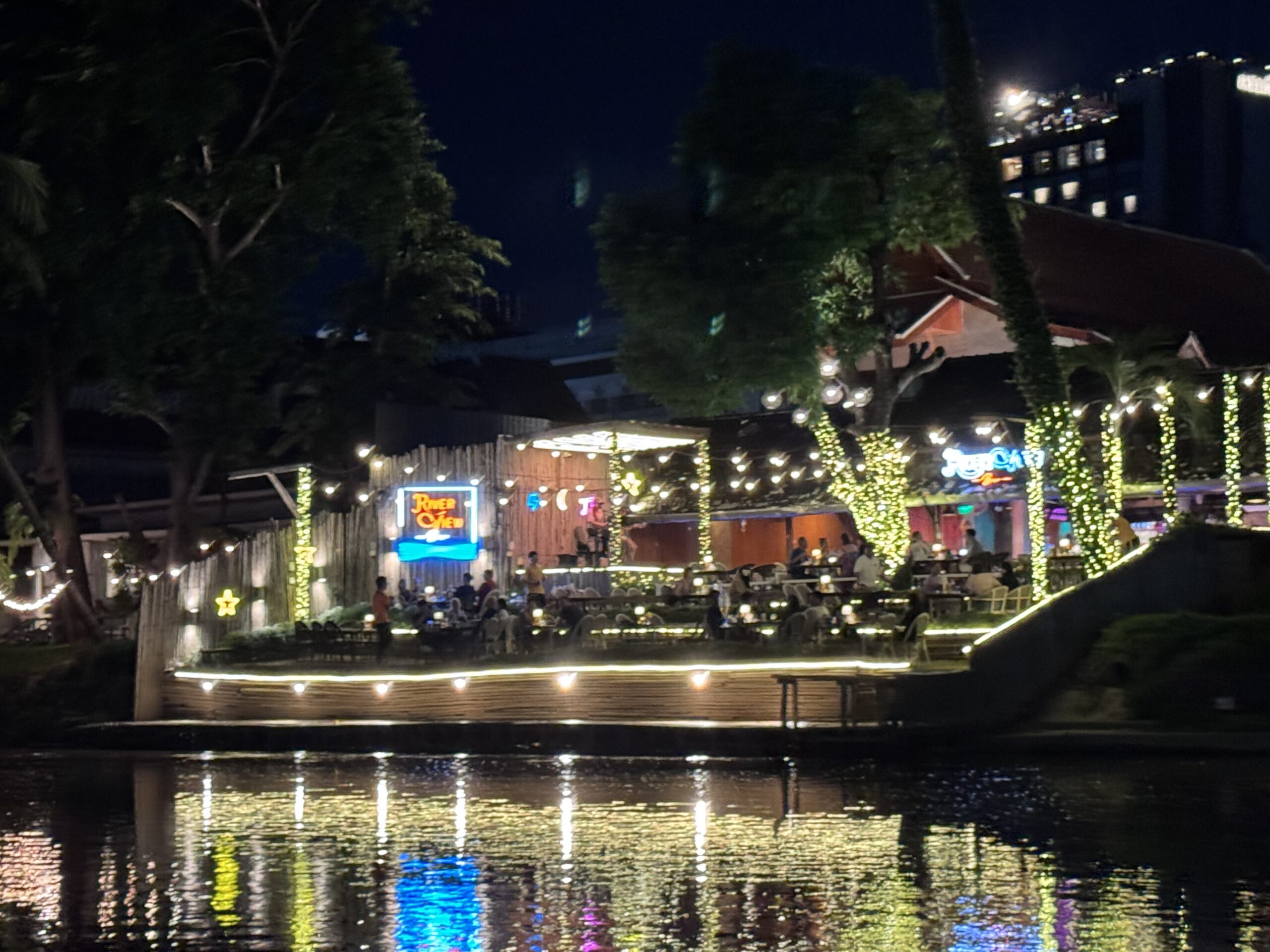 Lively riverside restaurant illuminated by colorful lights at night, with diners enjoying the ambiance and reflections shimmering on the water.