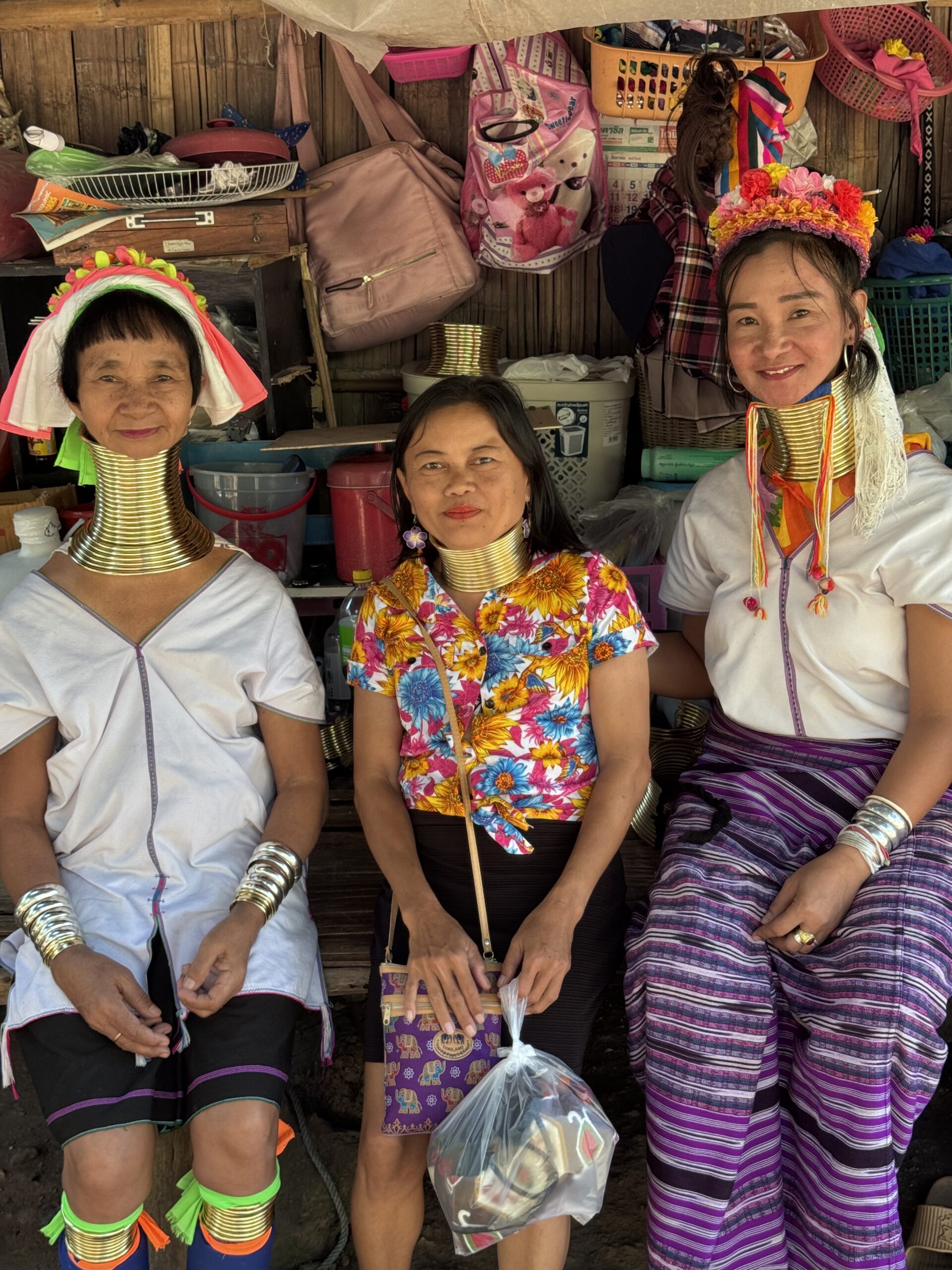 Three women from the Kayan Lahwi tribe showcase traditional brass neck coils and colorful clothing, with a backdrop of woven baskets and bags.