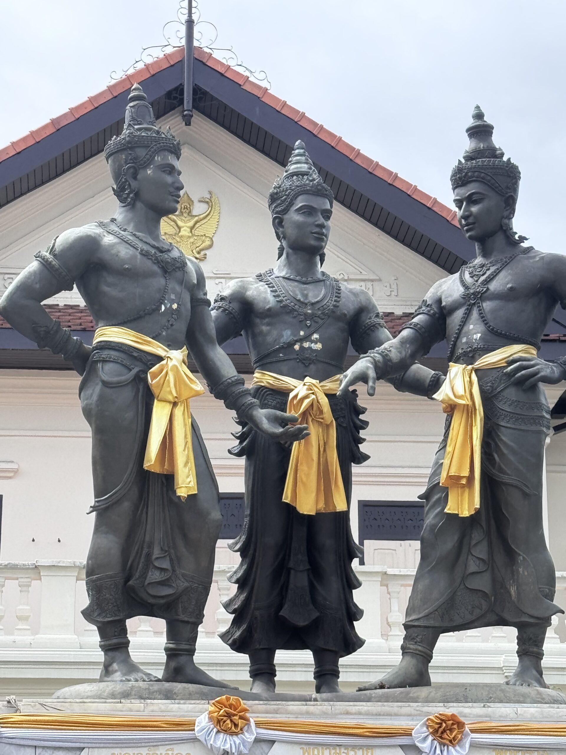 Three bronze statues depicting Hindu deities, wearing traditional attire and golden sashes, stand in front of a temple.