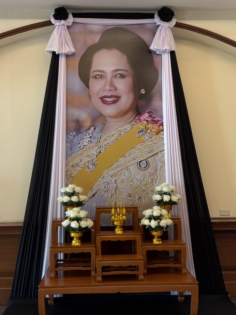 Portrait of a regal woman adorned in traditional attire, framed with black and white drapery, flanked by flower arrangements and ornamental stands.