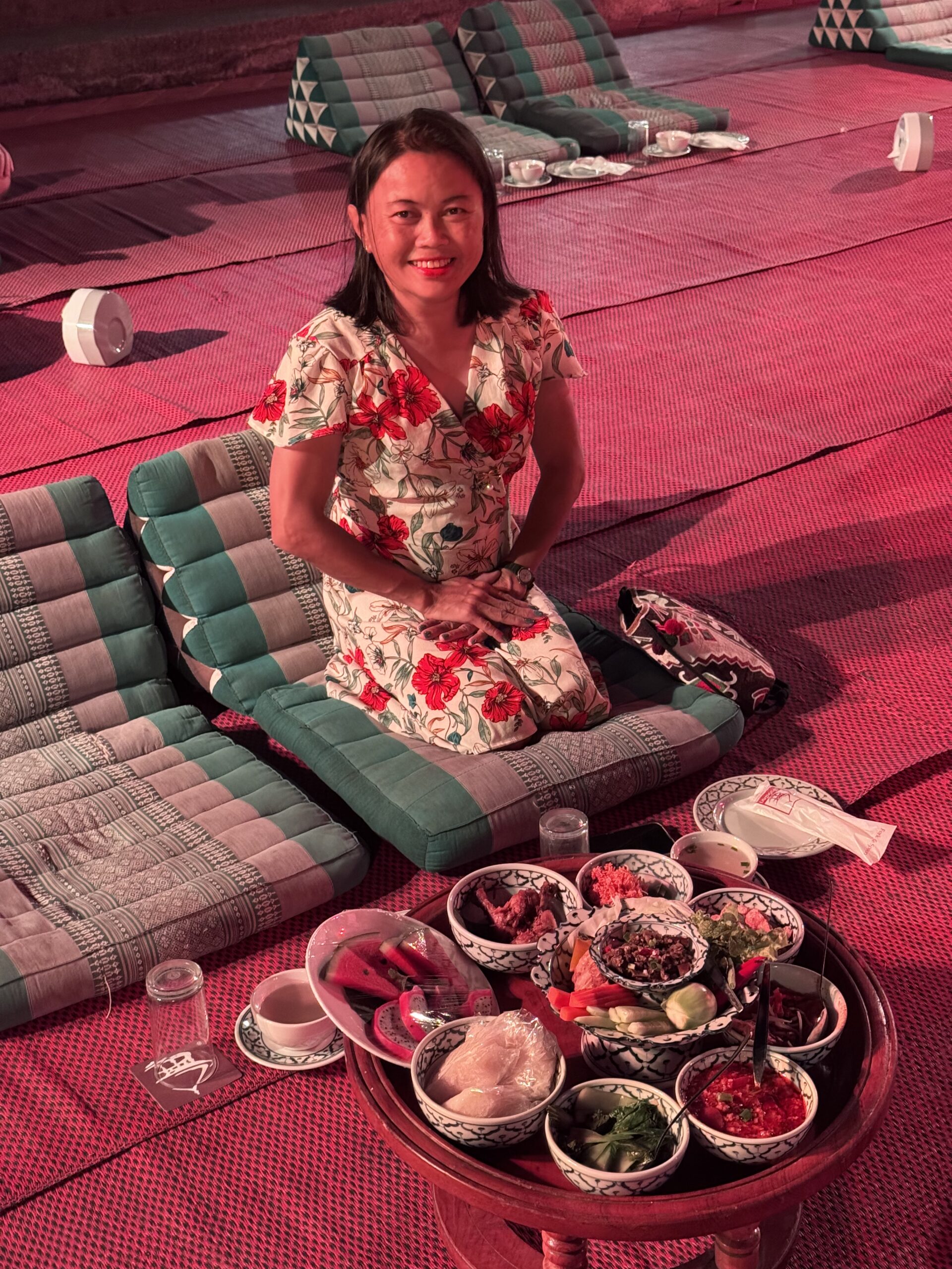 A woman sits on a mat surrounded by colorful cushions, posing with a table of assorted local dishes, showcasing a vibrant dining experience.