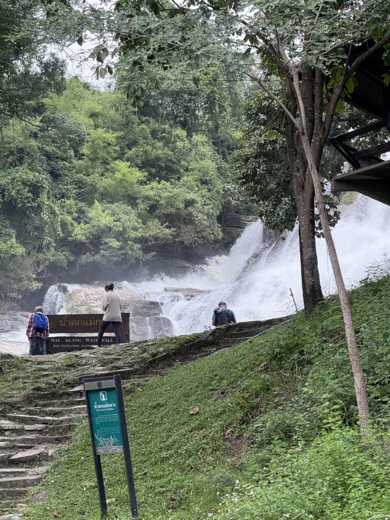 Visitors stand near Mae Klang Waterfall, surrounded by lush greenery. A sign identifies the location, highlighting its natural beauty.
